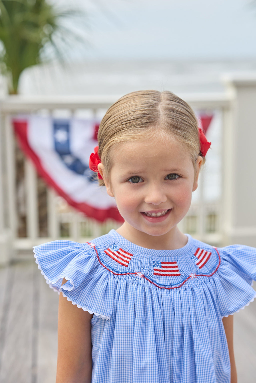 Little English traditional children's clothing. Flutter sleeve dress for toddler girls in royal blue mini gingham with smocking at the collar and American flag bunting