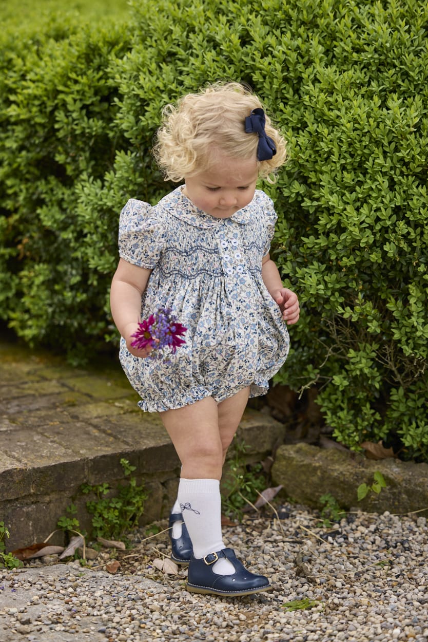 Little English classic children's clothing.  Dressy peter pan bubble for baby girls in blue Wisteria Floral with puff sleeves, ruffle details, and smocking at chest. Paired with white cotton knee high socks with navy bow embroidery