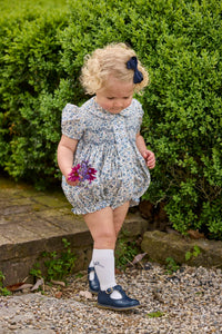 Little English classic children's clothing.  Dressy peter pan bubble for baby girls in blue Wisteria Floral with puff sleeves, ruffle details, and smocking at chest. Paired with white cotton knee high socks with navy bow embroidery