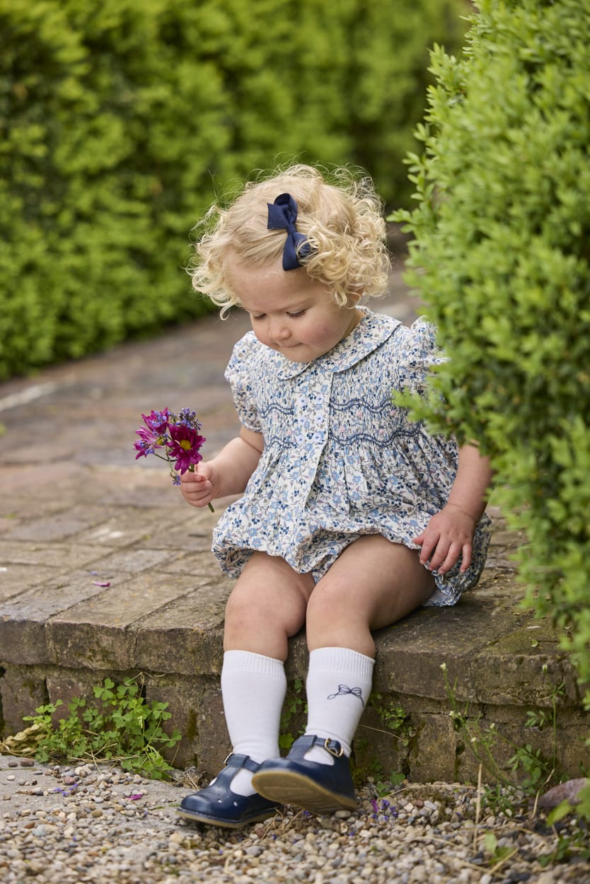 Little English classic children's clothing.  Dressy peter pan bubble for baby girls in blue Wisteria Floral with puff sleeves, ruffle details, and smocking at chest. Paired with white cotton knee high socks with navy bow embroidery