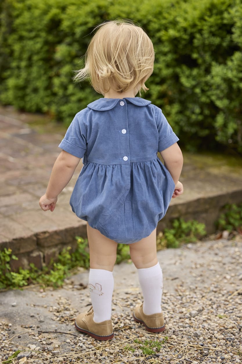 Little English classic children's clothing.  Baby boys stormy blue corduroy peter pan bubble with button tabs and flying mallard smocking across the chest. White knee high socks with mallard embroidery.