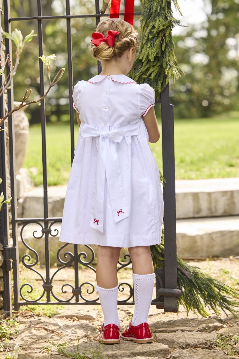 Little English traditional children's clothing.  Puff sleeve white peter pan dress with scalloped collar, red trim, and bow smocking across the chest.  Dressy outfit for little girls for the holiday