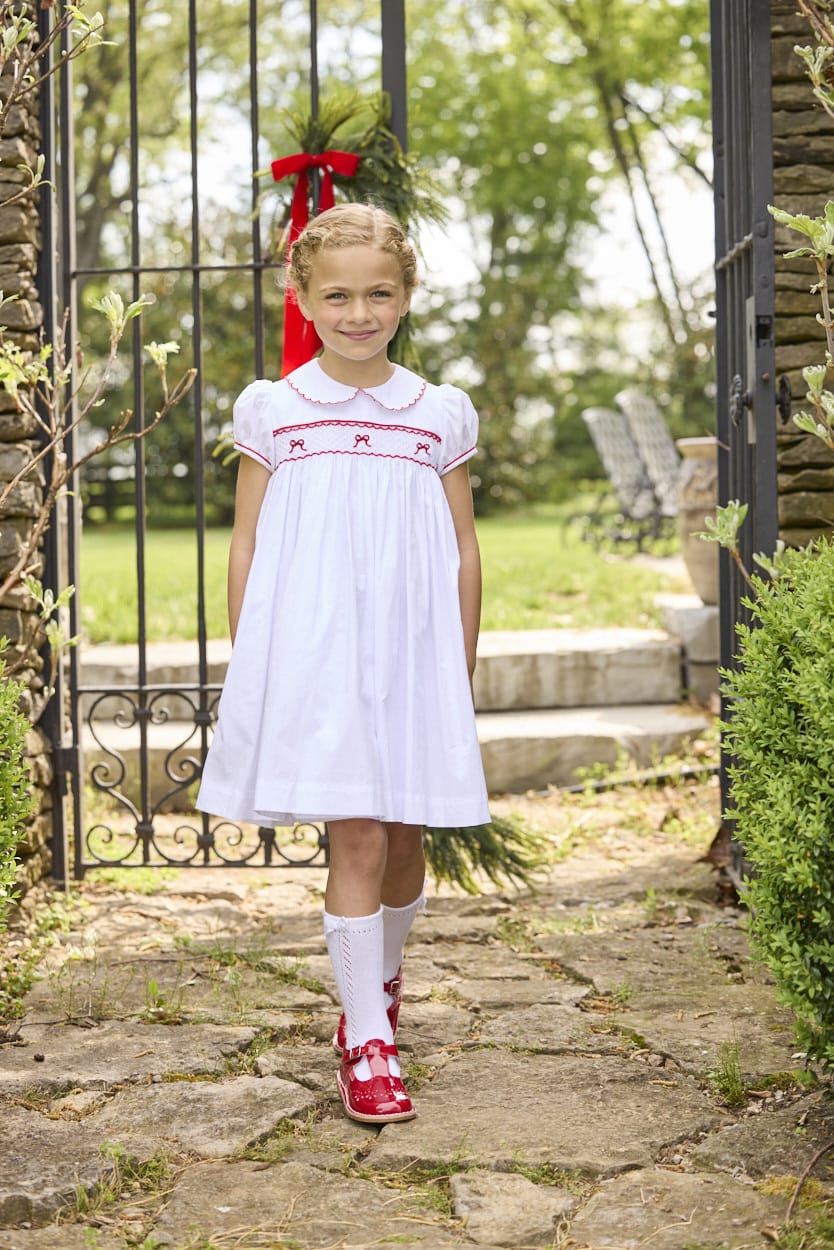 Little English traditional children's clothing.  Puff sleeve white peter pan dress with scalloped collar, red trim, and bow smocking across the chest.  Dressy outfit for little girls for the holiday