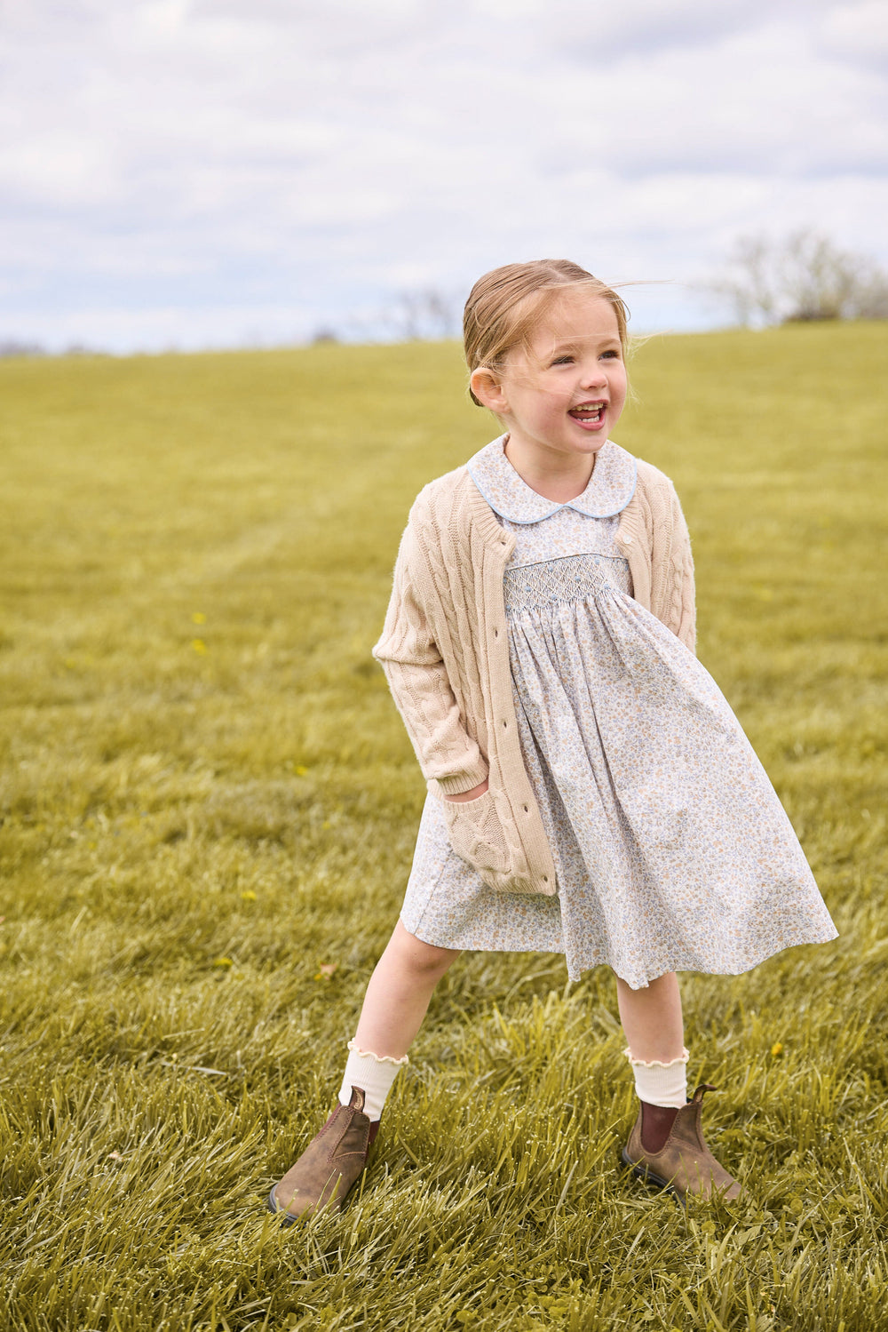 Front view of Smocked Peter Pan dress showing chest smocking and back sash, paired with oatmeal cardigan in grassy field