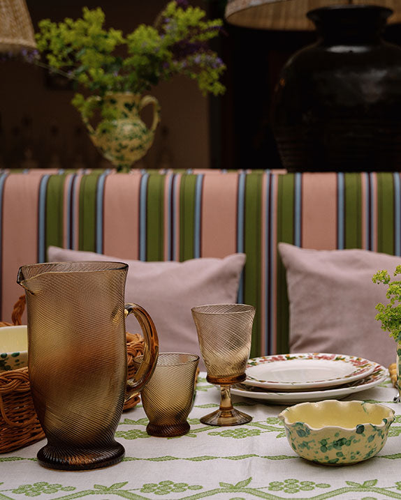 Chestnut brown spiral glass pitcher and tumblers on patterned tablecloth with stacked plates and striped banquette background