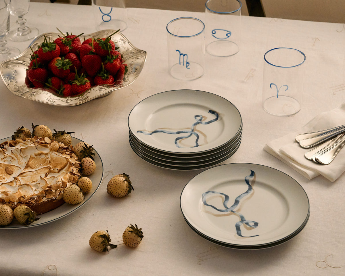  An elegantly set dining table with ribbon dessert plates in blue. There is a meringue pie topped with almonds and surrounded by small, champagne strawberries. To the left, a silver bowl full of ripe strawberries adds a pop of red color. The table is also adorned with several rocks glasses, each marked with different blue zodiac signs. Silverware is neatly placed on white napkins to the right. The tablecloth is white with embroidered details, providing a sophisticated setting for a holiday dinner party. 