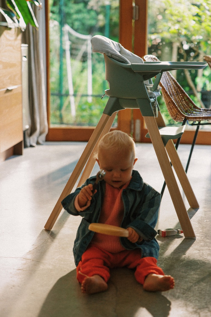 Baby playing on the floor beneath a Stokke Clikk high chair with Glacier Green cushion in a sunlit room