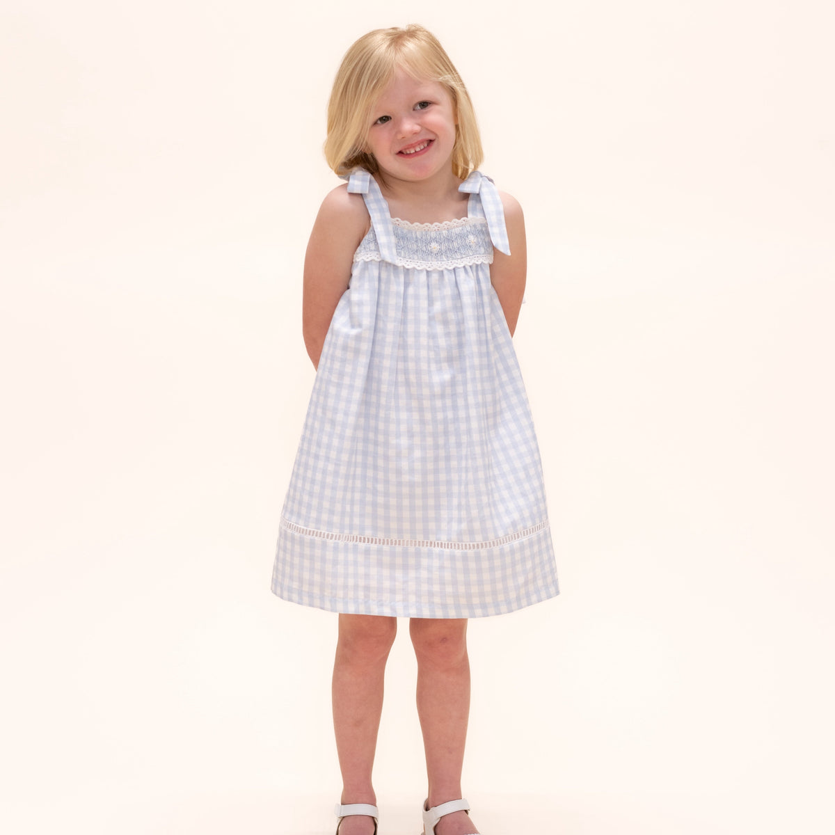 Front view of Gerbera Girl Dress showcasing blue and white gingham Spanish linen, cotton eyelet trim, and tie straps on a child
