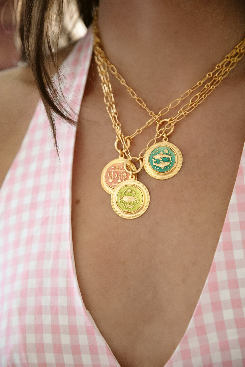 Close up of Sun Sign Necklace showing three layered gold zodiac coin pendants with colored enamel on paperclip chains worn over a pink gingham top