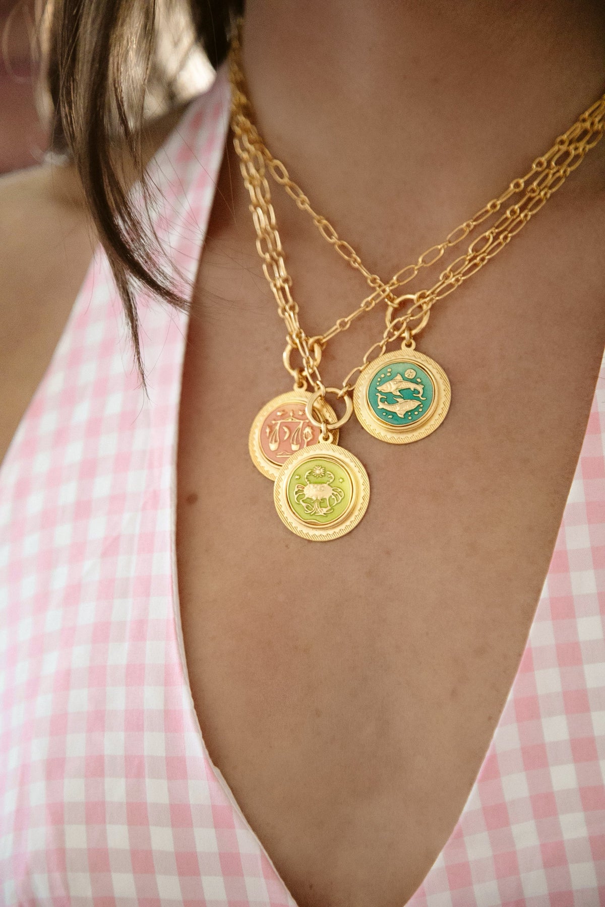Close up of Sun Sign Necklace showing three layered gold zodiac coin pendants with colored enamel on paperclip chains worn over a pink gingham top