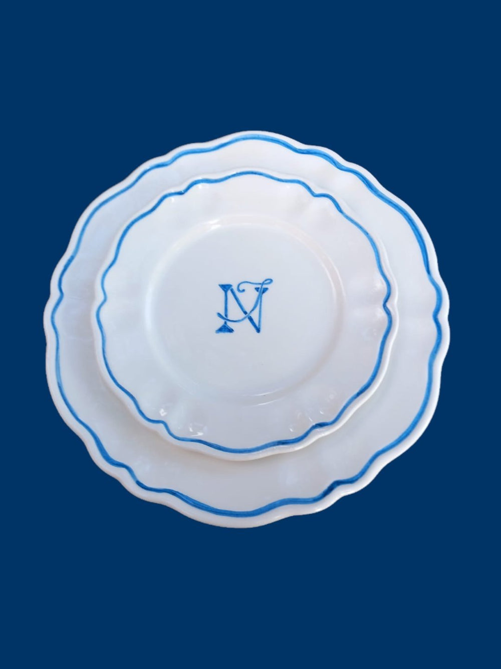 Top view of personalised scalloped plate set showing stacked white ceramic plates with blue hand-painted rim and monogram ‘N’ on navy background