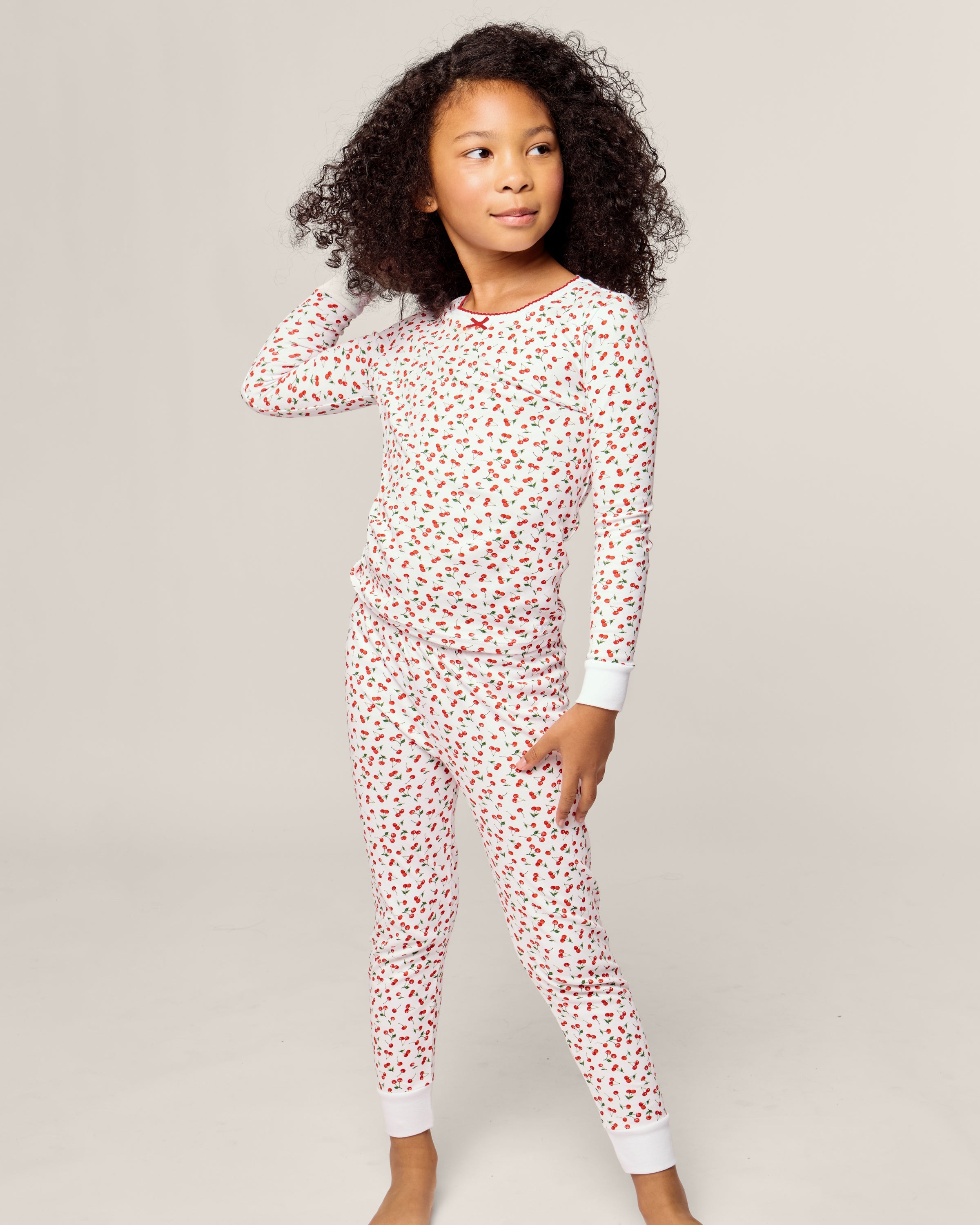 A child in Petite Plumes Kids Pima Snug Fit Pajama Set in Cherry Orchard stands barefoot on a plain backdrop. The white PJs with red hearts have white cuffs. With curly hair and relaxed posture, the child gazes off to the side, one hand resting on their head.