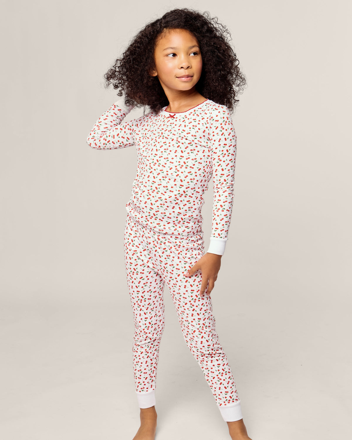 A child in Petite Plumes Kids Pima Snug Fit Pajama Set in Cherry Orchard stands barefoot on a plain backdrop. The white PJs with red hearts have white cuffs. With curly hair and relaxed posture, the child gazes off to the side, one hand resting on their head.