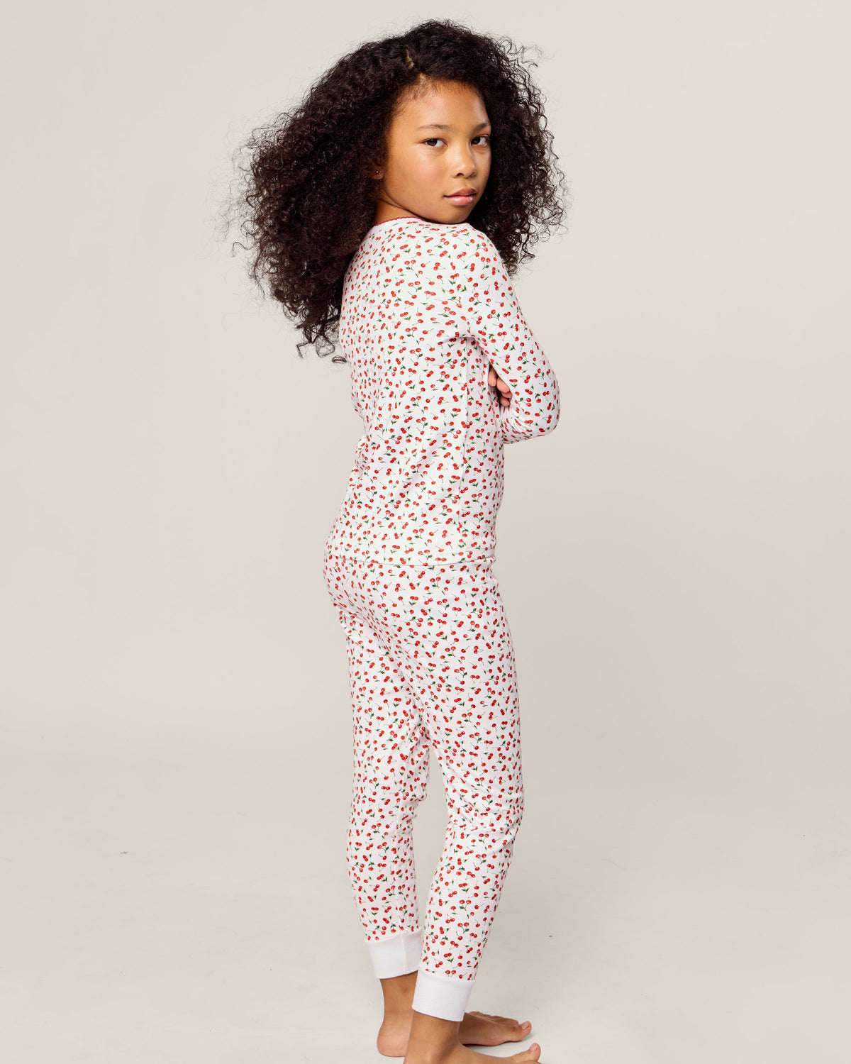 A child with curly hair stands sideways, barefoot in cozy Petite Plume Pima pajamas. The sleepwear, known as Kids Snug Fit Pajama Set in Cherry Orchard, features a cheerful red and black heart pattern. They cross their arms and gaze over their shoulder against a plain background.