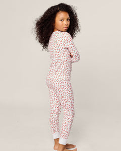 A child with curly hair stands sideways, barefoot in cozy Petite Plume Pima pajamas. The sleepwear, known as Kids Snug Fit Pajama Set in Cherry Orchard, features a cheerful red and black heart pattern. They cross their arms and gaze over their shoulder against a plain background.