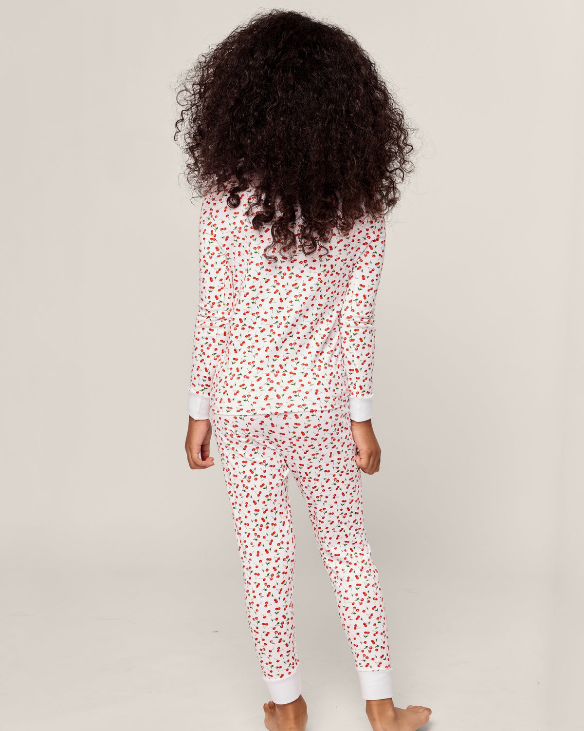 Dressed in Petite Plumes Kids Pima Snug Fit Pajama Set in Cherry Orchard, a curly-haired child stands barefoot on a light background. The soft pajamas, adorned with red hearts, envelop them as they face away from the camera, radiating warmth and comfort.