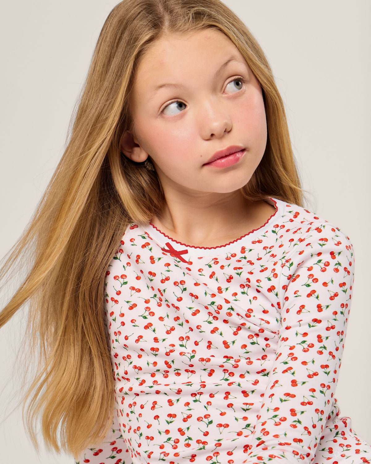 A young girl with long blonde hair, wearing the Petite Plume Kids Pima Snug Fit Pajama Set in Cherry Orchard print, looks to the side thoughtfully against a plain, neutral background.