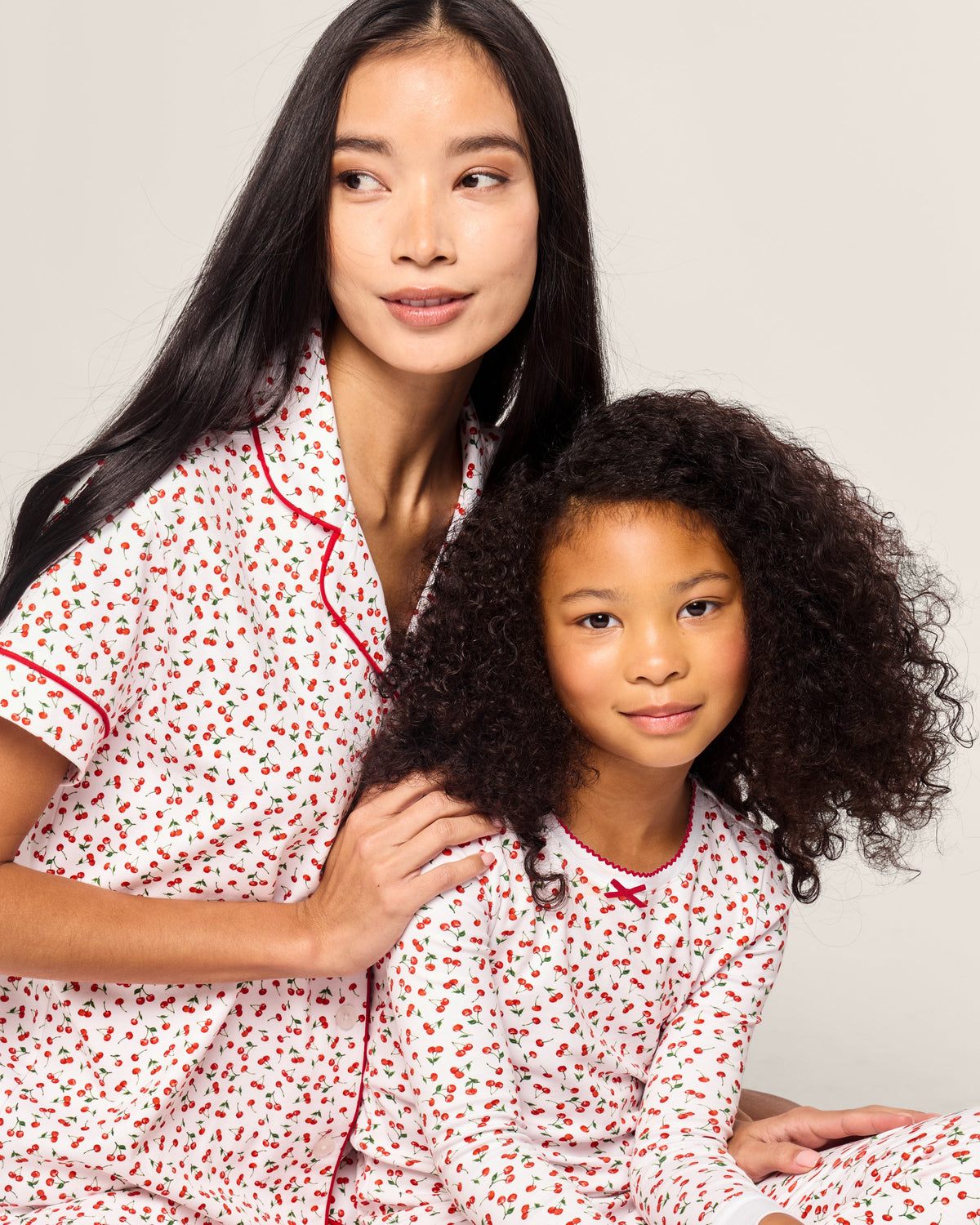 A woman with long black hair and a child with curly hair wear matching Petite Plume sleepwear, featuring the Kids Pima Snug Fit Pajama Set in Cherry Orchard. They sit closely together, looking to the side with gentle smiles.