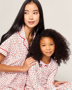 A woman with long black hair and a child with curly hair wear matching Petite Plume sleepwear, featuring the Kids Pima Snug Fit Pajama Set in Cherry Orchard. They sit closely together, looking to the side with gentle smiles.