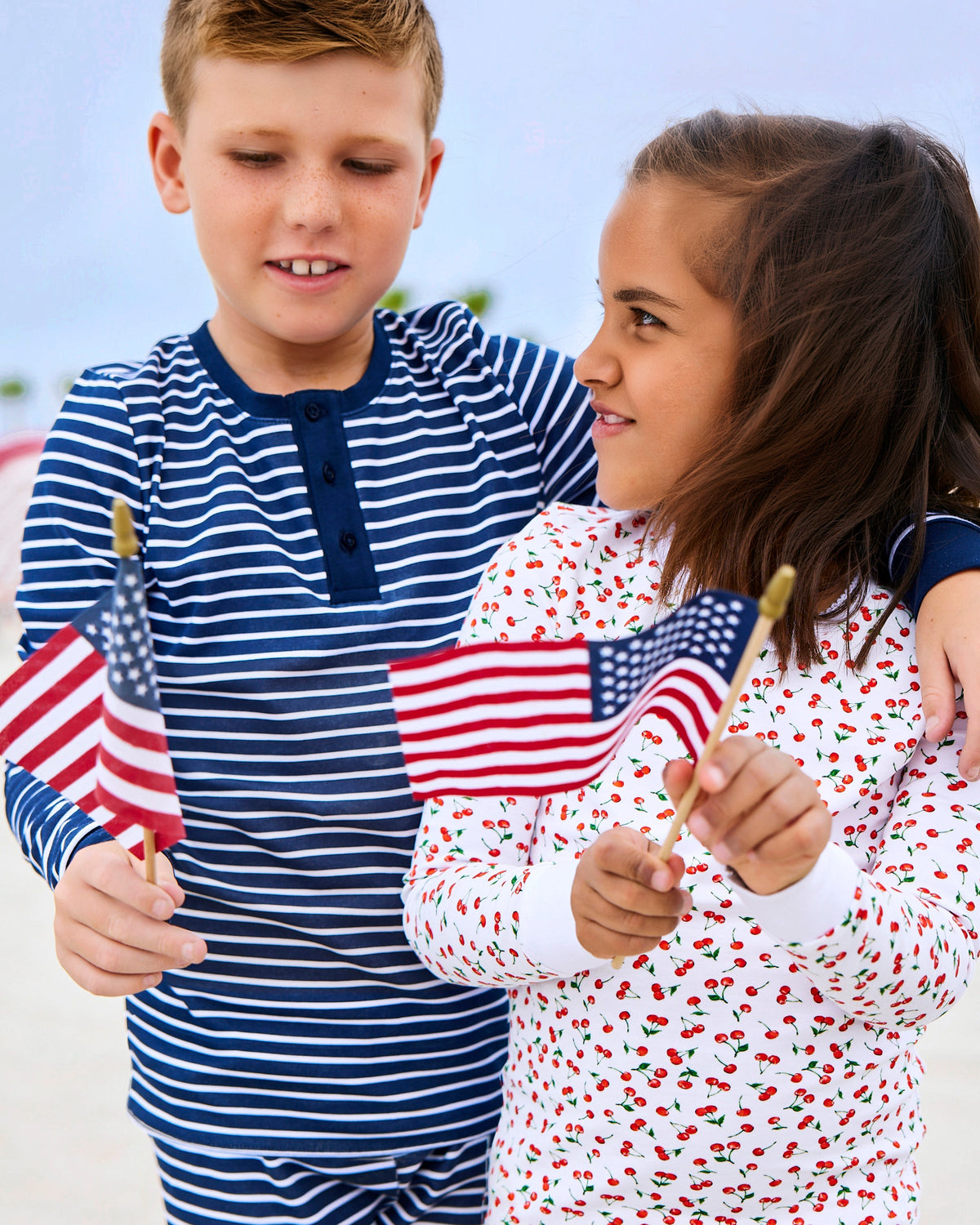 In Petite Plume attire, a boy in a blue and white striped shirt and a girl in the Kids Pima Snug Fit Pajama Set in Cherry Orchard embrace coordinated style, holding American flags. They stand outdoors with a blurred backdrop, showcasing chic Mom-and-me fashion.