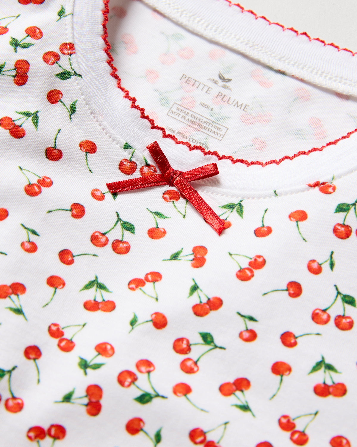 The Kids Pima Snug Fit Pajama Set in Cherry Orchard by Petite Plume features a close-up of the white shirt in luxurious Pima Cotton, adorned with small red cherries and green stems. A tiny red bow decorates the neckline, and decorative red stitching lines the collar edge.
