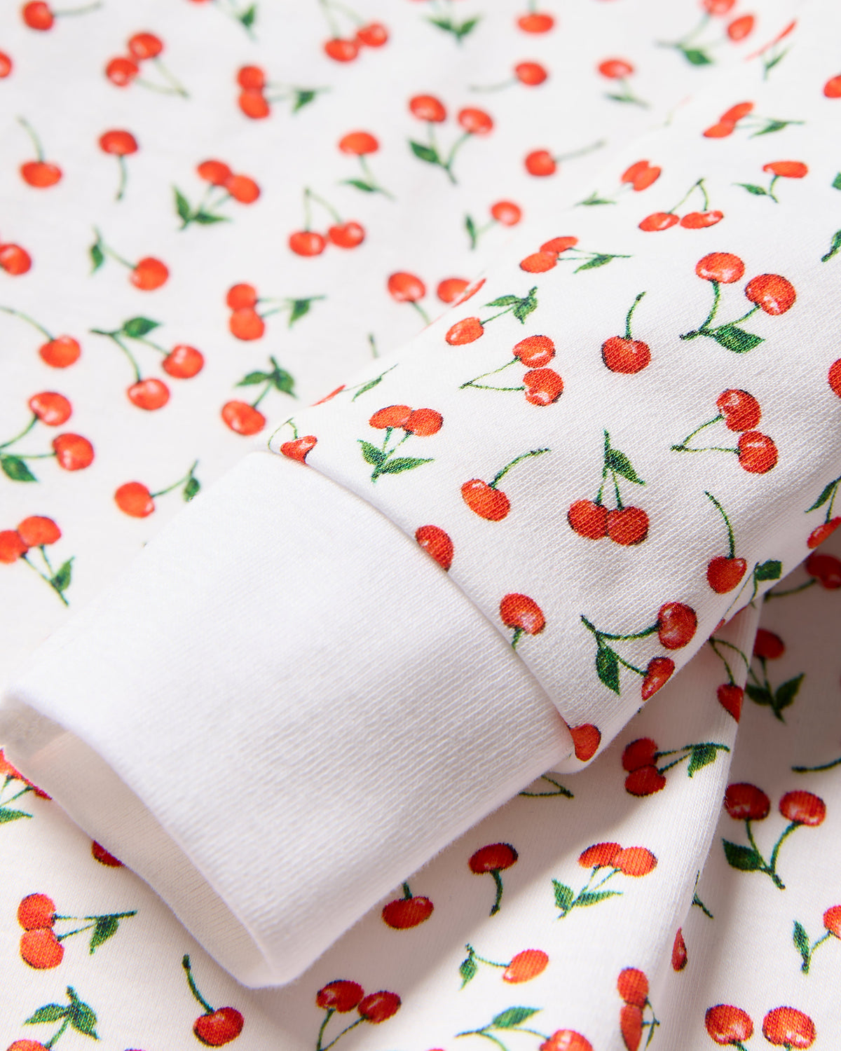 Close-up of white fabric with a cherry print, ideal for coordinated family sleepwear. The Kids Pima Snug Fit Pajama Set in Cherry Orchard by Petite Plume features bright red cherries with green leaves on cotton material. A folded sleeve edge displays the same design consistently.