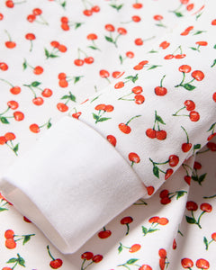Close-up of white fabric with a cherry print, ideal for coordinated family sleepwear. The Kids Pima Snug Fit Pajama Set in Cherry Orchard by Petite Plume features bright red cherries with green leaves on cotton material. A folded sleeve edge displays the same design consistently.