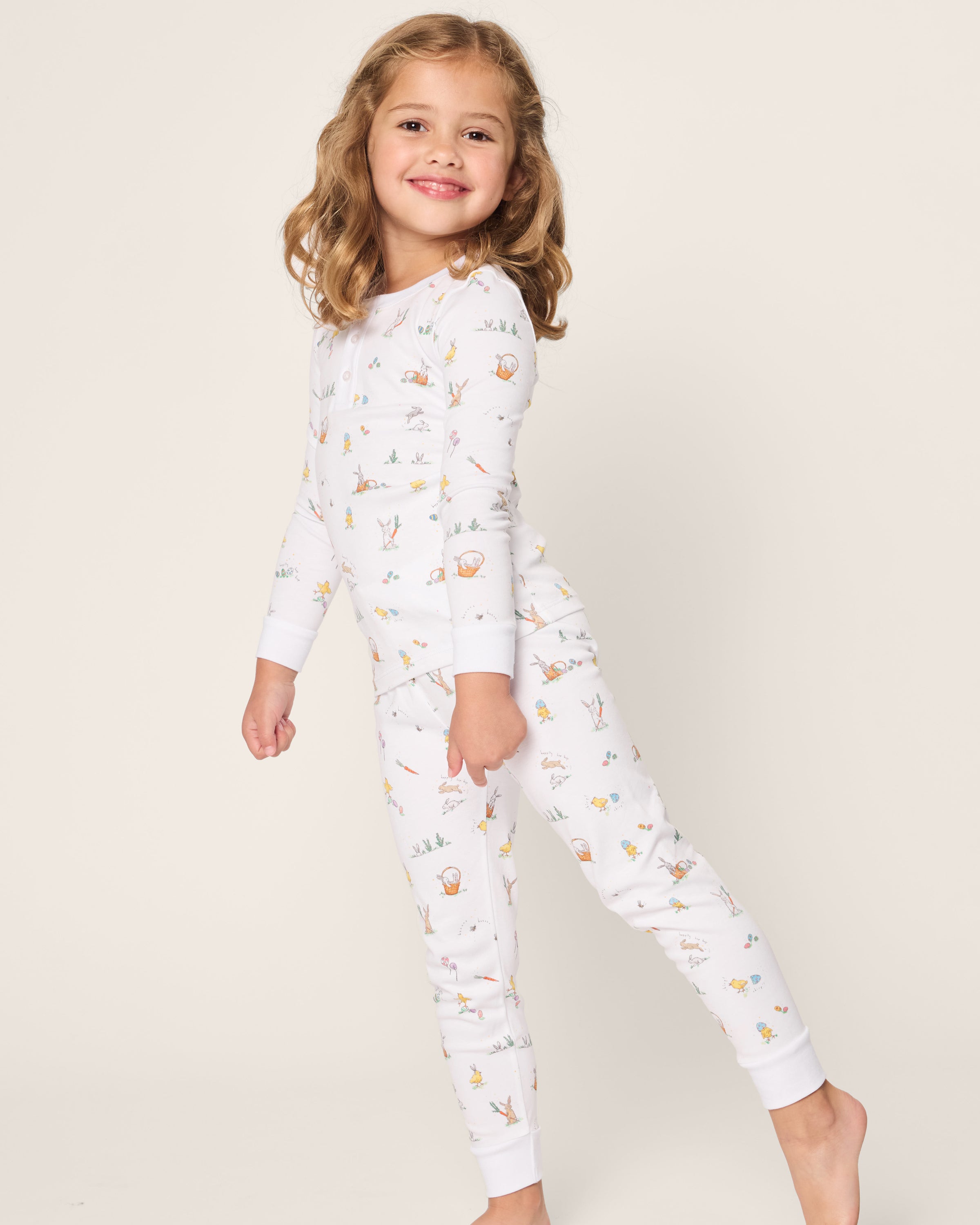 A young girl with light brown hair smiles sideways, showcasing her style in the Petite Plume Kids Pima Snug Fit Pajama Set in Easter Frolic, featuring colorful animal prints. Made from soft Pima cotton, it ensures comfort as she plays against a plain off-white background.