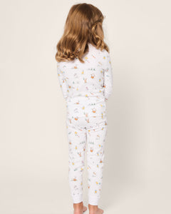 A child with wavy brown hair stands barefoot, facing away from the camera in their white Petite Plume Kids Pima Snug Fit Pajama Set in Easter Frolic, featuring small animal and flower illustrations on a light beige background.