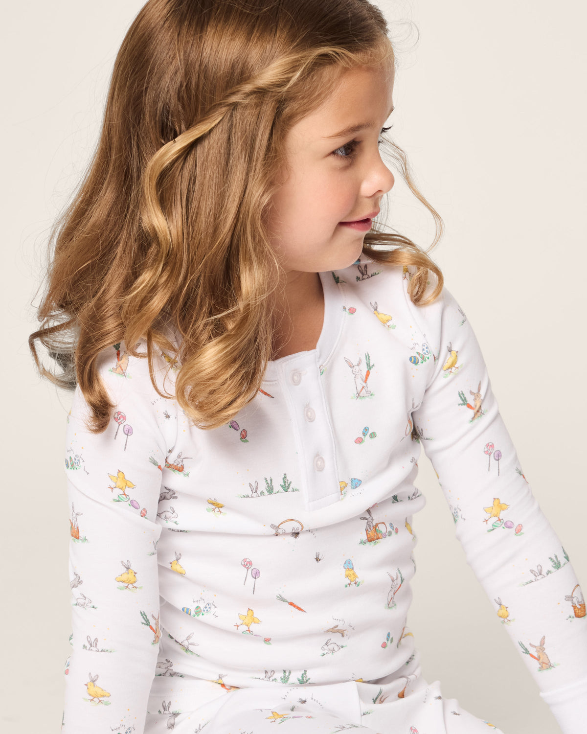 A child with curly brown hair, dressed in Petite Plumes Kids Pima Snug Fit Pajama Set in Easter Frolic, sits against a light background. She gazes to her right with a gentle expression, showcasing her coordinated style and embodying the playful essence of Easter Frolic.