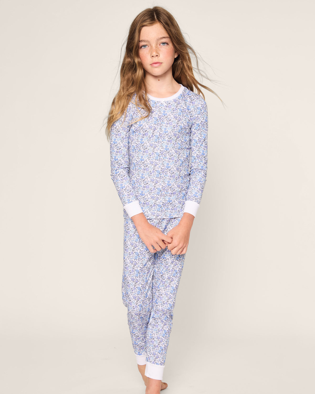 A young girl with long brown hair stands barefoot against a light background, dressed in Petite Plumes Kids Pima Snug Fit Pajama Set in Fleur dAzur, featuring a blue and white vintage-inspired floral print. She looks directly at the camera with a neutral expression.