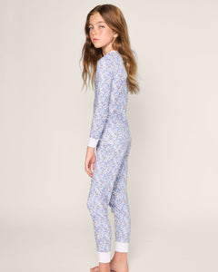 A young girl with long brown hair stands sideways, looking at the camera. She wears Petite Plumes Kids Pima Snug Fit Pajama Set in Fleur dAzur, featuring a cozy long sleeve top and pants made from soft Pima Cotton with a vintage-inspired blue and white floral print against a plain background.