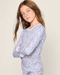 A young girl with long brown hair poses confidently, arms crossed, in Petite Plumes Kids Pima Snug Fit Pajama Set in Fleur dAzur. The vintage-inspired floral-patterned outfit crafted from soft Pima cotton stands out against the neutral background with timeless charm.