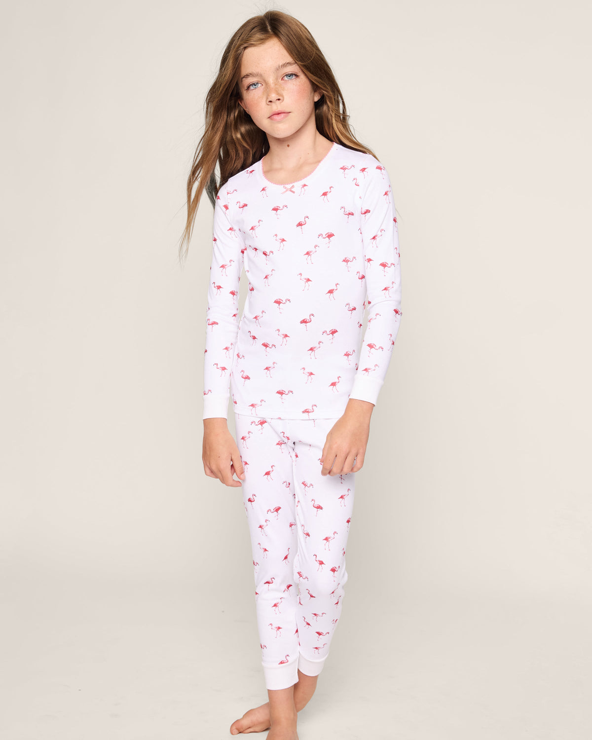 A young girl with long brown hair stands barefoot in a child’s wardrobe against a plain, light background, wearing the cozy Petite Plume Kids Pima Snug Fit Pajama Set in Flamingo featuring a red flamingo pattern on her long sleeve top and pant set.