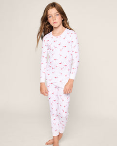 A young girl with long brown hair stands barefoot in a child’s wardrobe against a plain, light background, wearing the cozy Petite Plume Kids Pima Snug Fit Pajama Set in Flamingo featuring a red flamingo pattern on her long sleeve top and pant set.