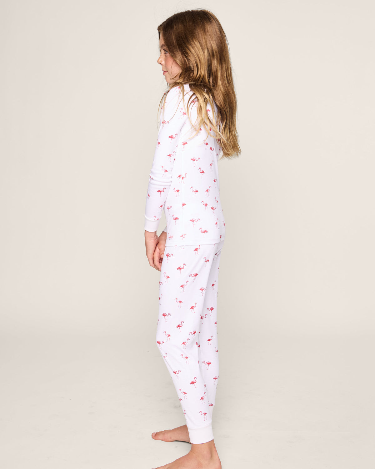A young girl with long brown hair stands barefoot in profile, wearing Petite Plumes Kids Pima Snug Fit Pajama Set in Flamingo—a cozy white sleepwear ensemble with small pink flamingo prints. The stylish long sleeve top and pants are set against a plain, light-colored background.