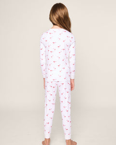 A barefoot child with long hair stands facing away, wearing the Petite Plume Kids Pima Snug Fit Pajama Set in Flamingo. The cozy long sleeve top and pant set, made from soft Pima cotton sleepwear, features a charming pink flamingo pattern on a light-colored background.