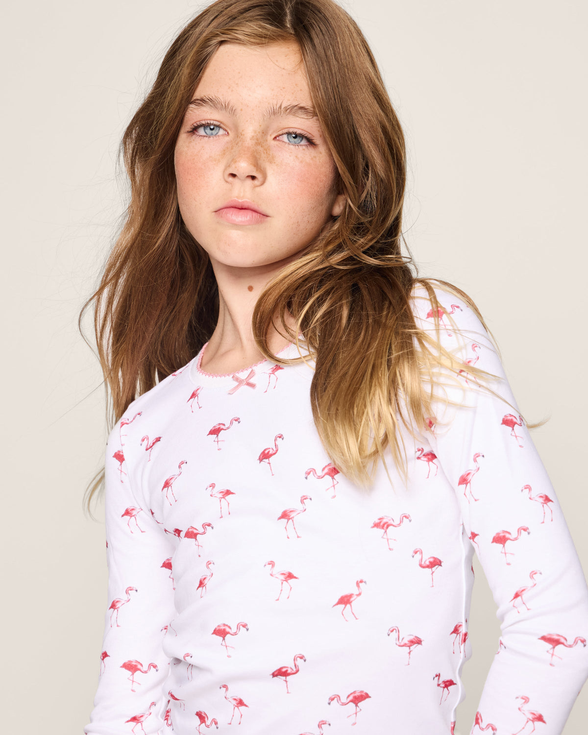 A young girl with long brown hair wears the Kids Pima Snug Fit Pajama Set in Flamingo by Petite Plume. Her neutral expression contrasts with the vibrant flamingo design against a light background, embodying cool comfort and style in her childs wardrobe.