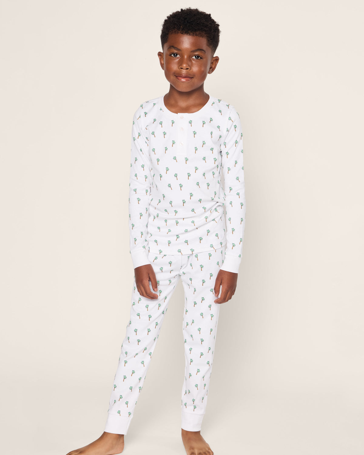A child is happily dressed in the Kids Pima Snug Fit Pajama Set in Palmier by Petite Plume, featuring a matching white long sleeve top and bottoms with a green dinosaur print. Made from soft Pima cotton, they stand barefoot against a plain background with hands by their sides.