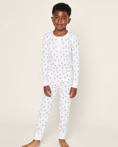 A child is happily dressed in the Kids Pima Snug Fit Pajama Set in Palmier by Petite Plume, featuring a matching white long sleeve top and bottoms with a green dinosaur print. Made from soft Pima cotton, they stand barefoot against a plain background with hands by their sides.