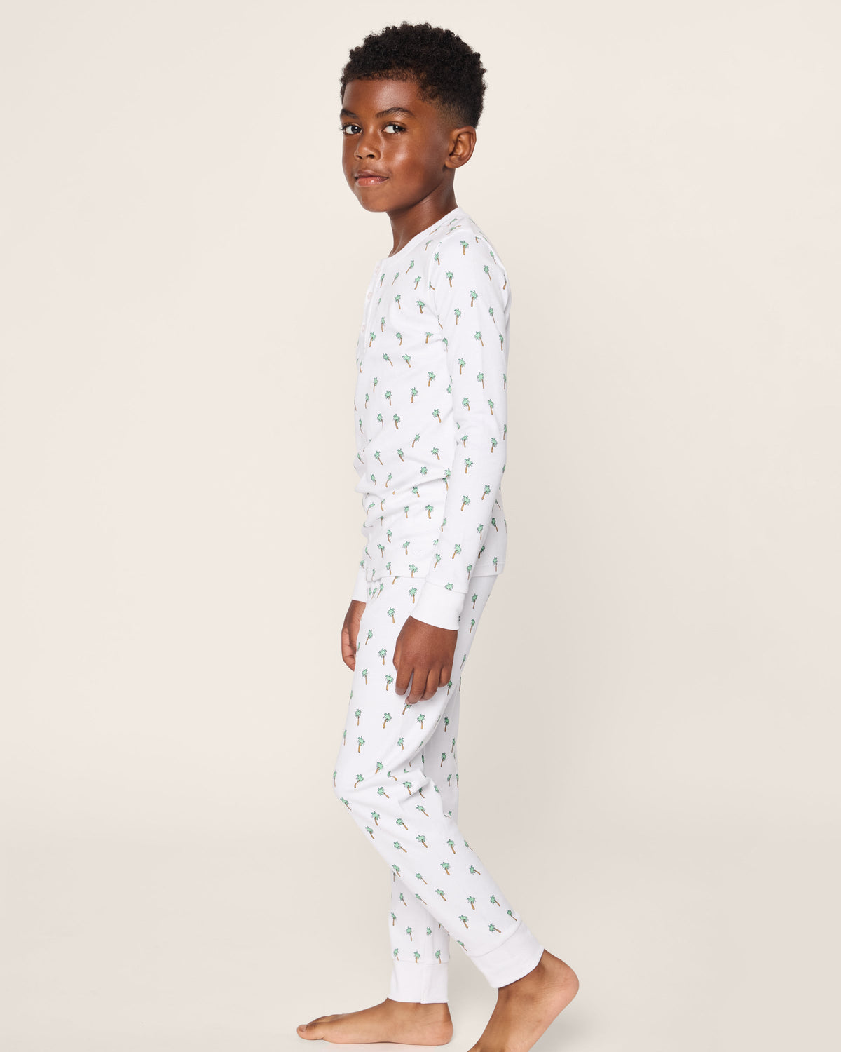 A young boy stands sideways on a white background, barefoot in Petite Plume’s Kids Pima Snug Fit Pajama Set in Palmier. The luxurious sleepwear features a cozy long sleeve top with a soft Pima cotton design adorned with small green cacti.