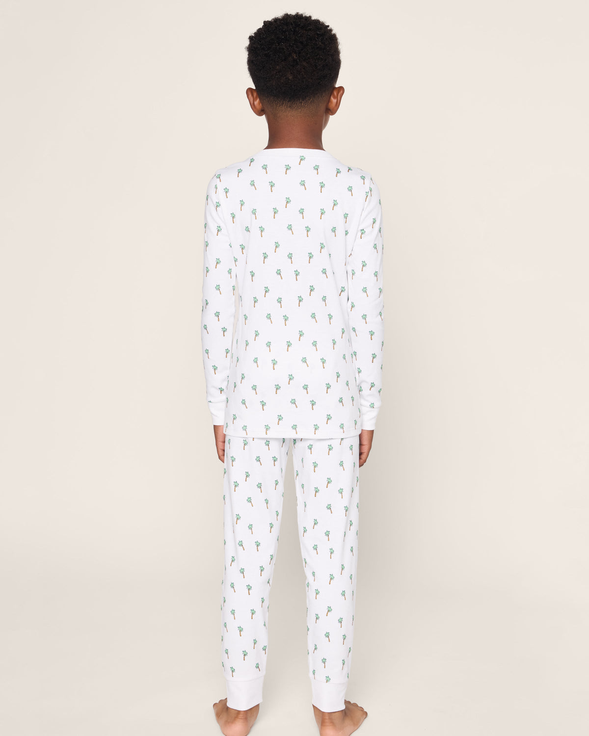 A barefoot child with short curly hair wears Petite Plumes luxurious Kids Pima Snug Fit Pajama Set in Palmier. The white sleepwear, featuring a green and blue floral pattern, includes a long sleeve top against an off-white background.