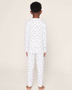 A barefoot child with short curly hair wears Petite Plumes luxurious Kids Pima Snug Fit Pajama Set in Palmier. The white sleepwear, featuring a green and blue floral pattern, includes a long sleeve top against an off-white background.