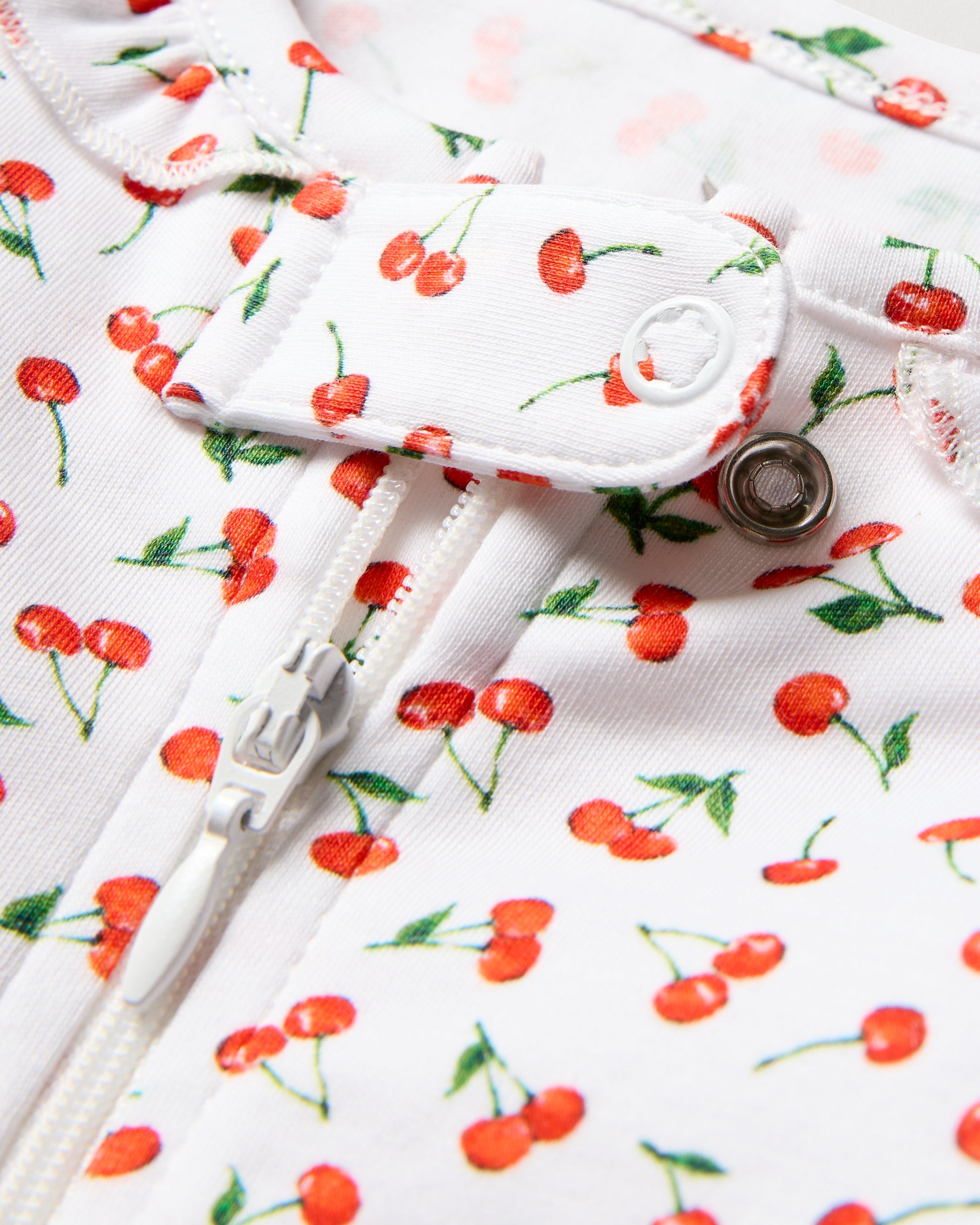 Close-up of Petite Plumes Baby Pima Romper in Cherry Orchard print, made from soft Pima cotton with a zipper and snap button. The playful design features small red cherries with green stems, evenly scattered for added charm.