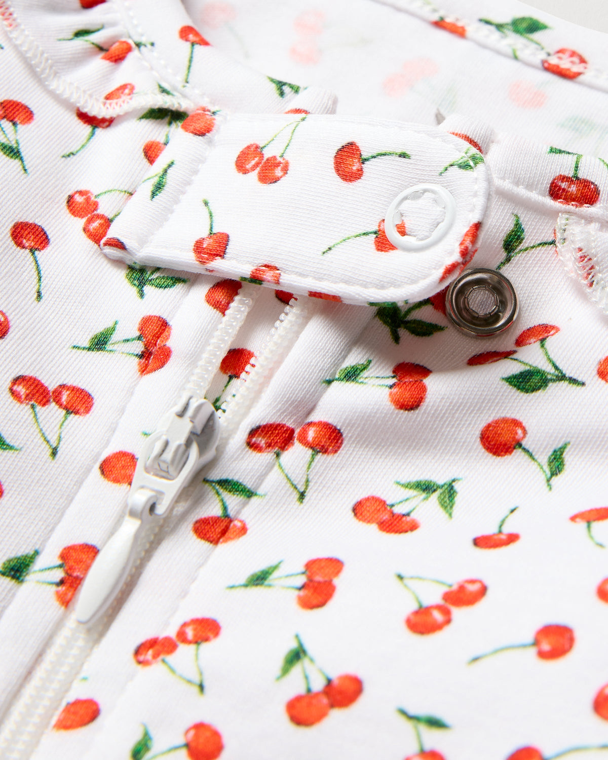 Close-up of Petite Plumes Baby Pima Romper in Cherry Orchard print, made from soft Pima cotton with a zipper and snap button. The playful design features small red cherries with green stems, evenly scattered for added charm.