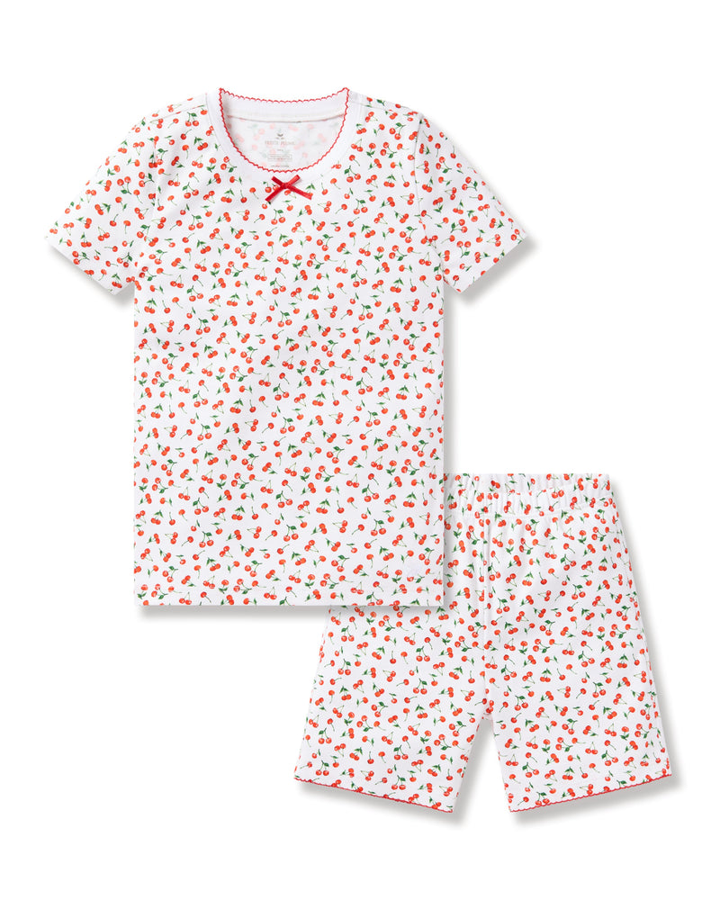 The Petite Plume Kids Pima Snug Fit Short Set in Cherry Orchard features a luxurious Pima Cotton design with a white base, playful red strawberry and green leaf pattern, stylish silhouette, red bow at the neckline, and matching red trim on the short-sleeved top and shorts.