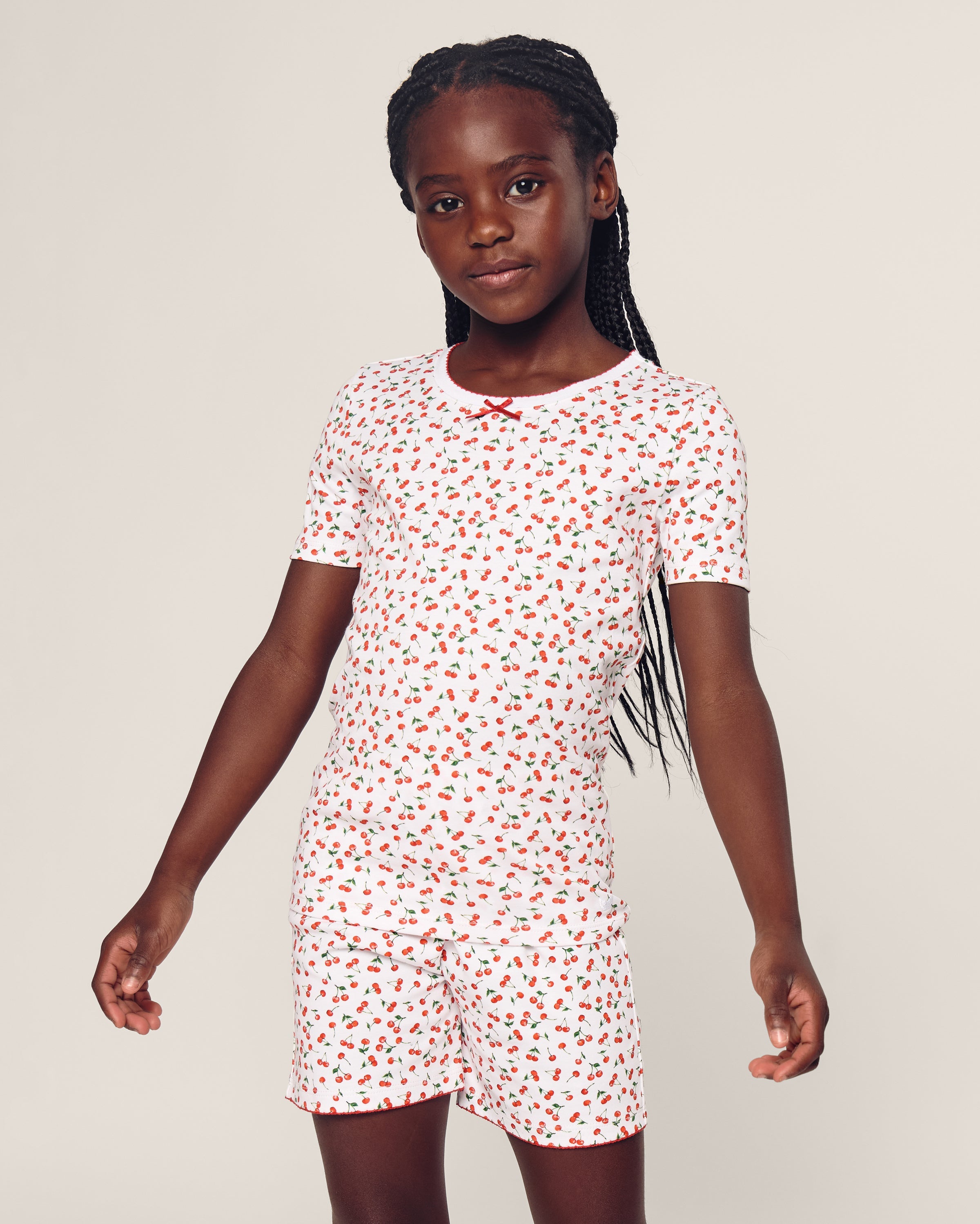 A young girl in Petite Plumes Kids Pima Snug Fit Short Set in Cherry Orchard print stands confidently against a neutral background, her long braided hair cascading over her shoulder. The stylish silhouette enhances the vibrant pattern, adding charm to her look.