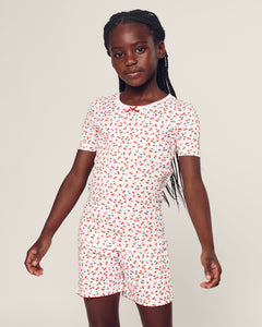 A young girl in Petite Plumes Kids Pima Snug Fit Short Set in Cherry Orchard print stands confidently against a neutral background, her long braided hair cascading over her shoulder. The stylish silhouette enhances the vibrant pattern, adding charm to her look.