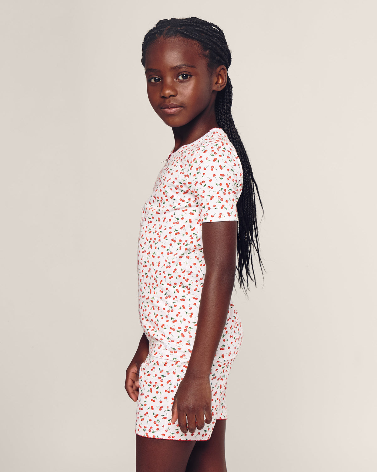 A young girl with long braided hair wears the Petite Plume Kids Pima Snug Fit Short Set in Cherry Orchard, standing in profile against a plain background, showcasing a stylish silhouette.