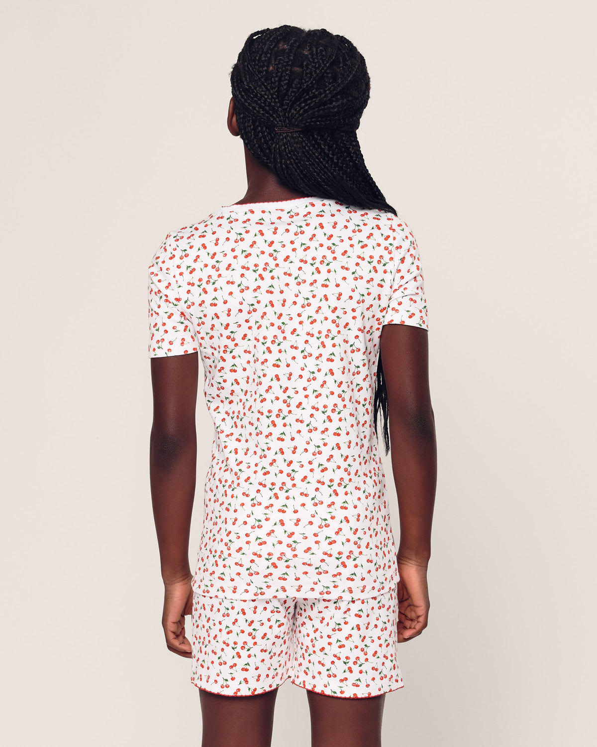 A person with braided hair is dressed in the elegant Kids Pima Snug Fit Short Set in Cherry Orchard by Petite Plume, featuring a white t-shirt and shorts adorned with a small red flower print. The plain off-white background enhances the stylish silhouette of the outfit.